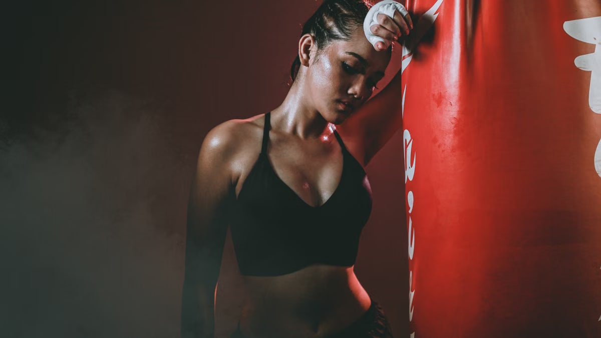 An athletic woman in a boxing gym taking a break beside a punching bag. Captured in moody lighting.